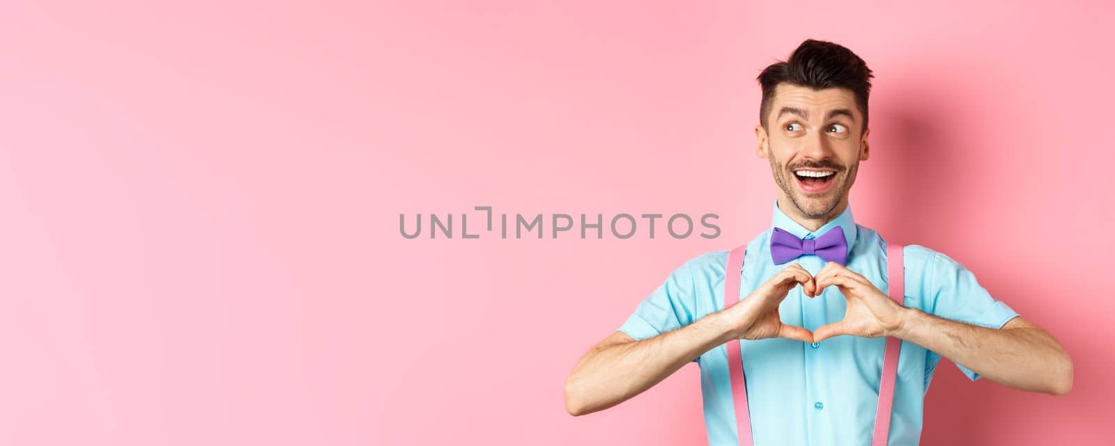 Happy Valentines day. Cheerful male model enjoying romantic date, showing heart sign, say I love you and looking left with happy smile, standing over pink background.