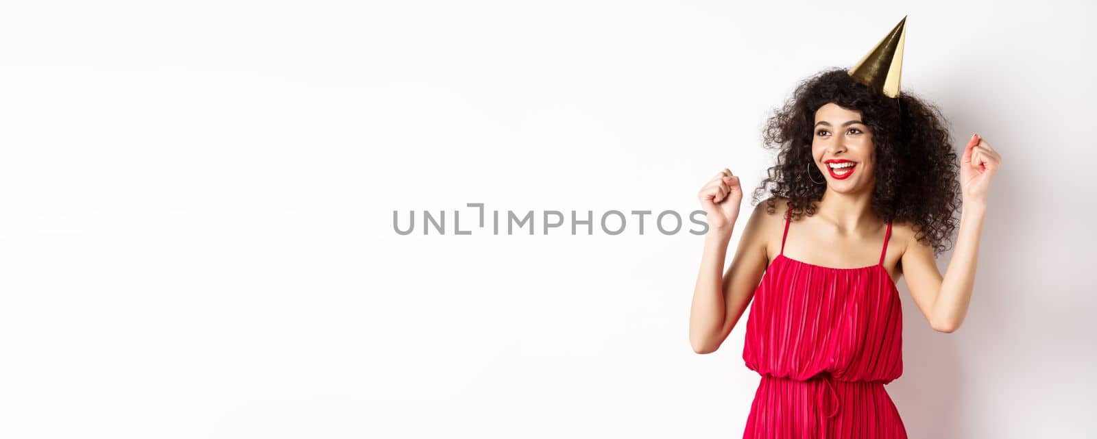 Happy birthday girl celebrating, wearing party hat and red dress, dancing and having fun, standing against white background by Benzoix