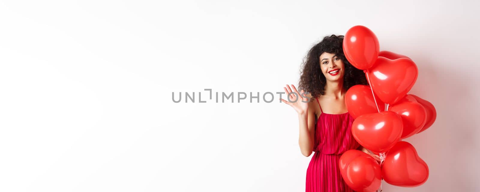 Beautiful lady with curly hair, standing near valentines holiday balloons and waving hand, saying hello, standing against white background.