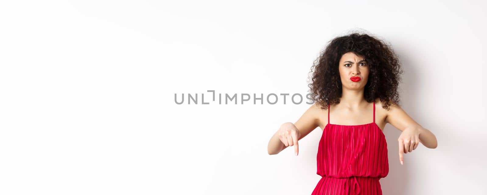 Disappointed caucasian woman in red dress complaining on spoiled date, frowning and grimacing, standing over white background.