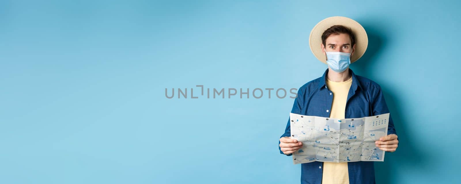 Covid-19, pandemic and travel concept. Happy tourist on summer vacation wearing medical mask and straw hat, holding road map, standing on blue background.