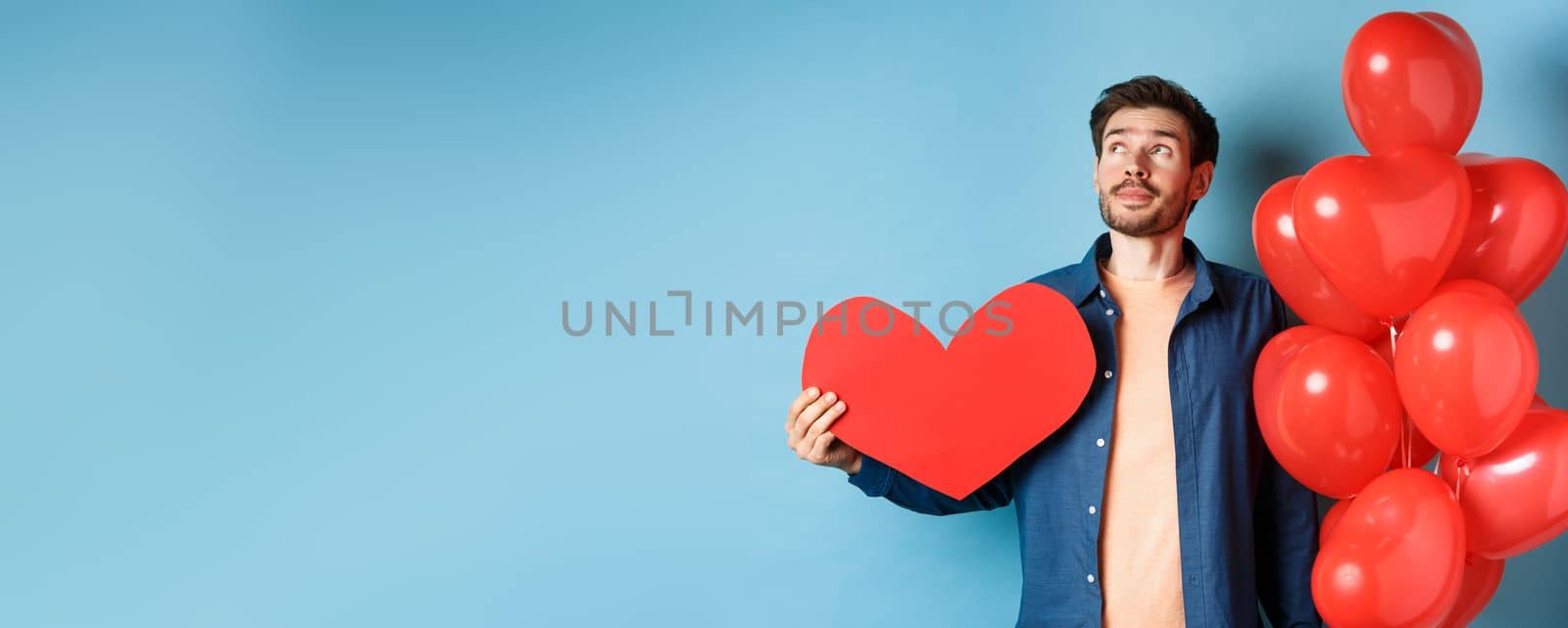 Valentines day and love concept. Man dreaming of soulmate, holding big red heart cutout and balloons, standing over blue background.