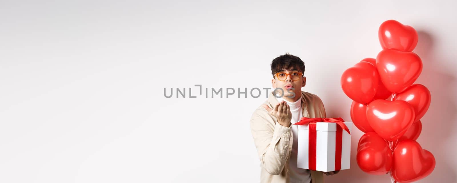 Valentines day and romance concept. Romantic modern man holding special gift for lover and sending air kiss at camera, standing near hearts balloons, white background.
