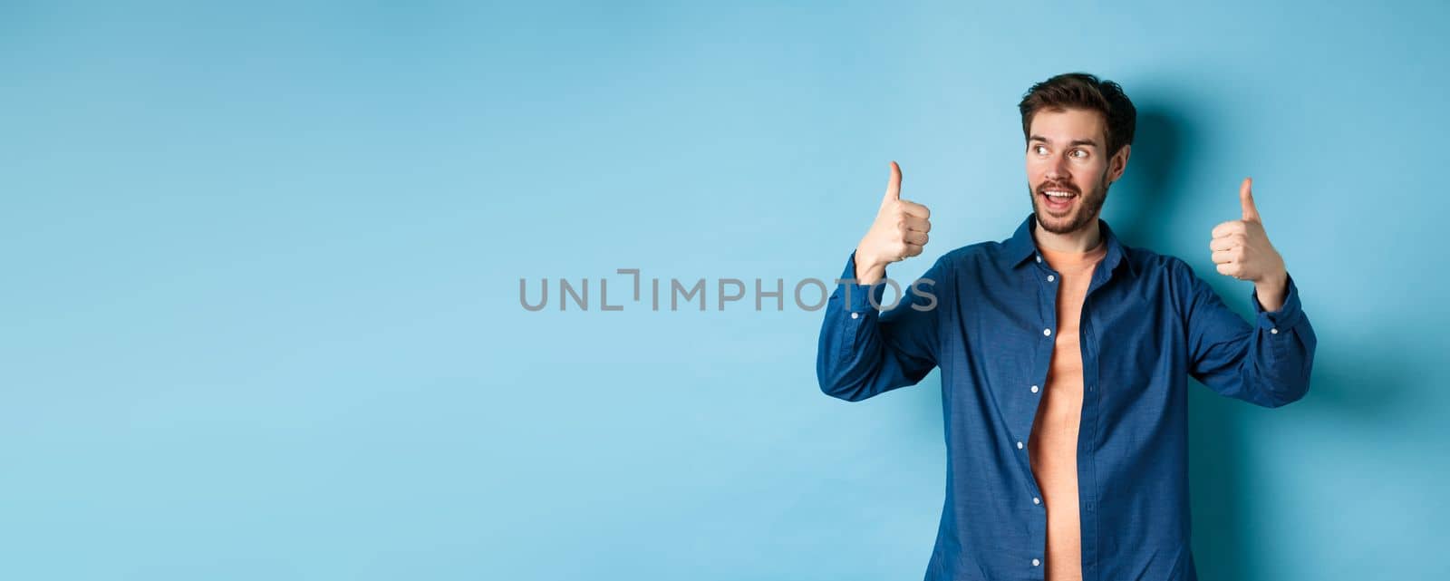 Handsome young man looking aside at empty space and showing thumbs up, standing on blue background.