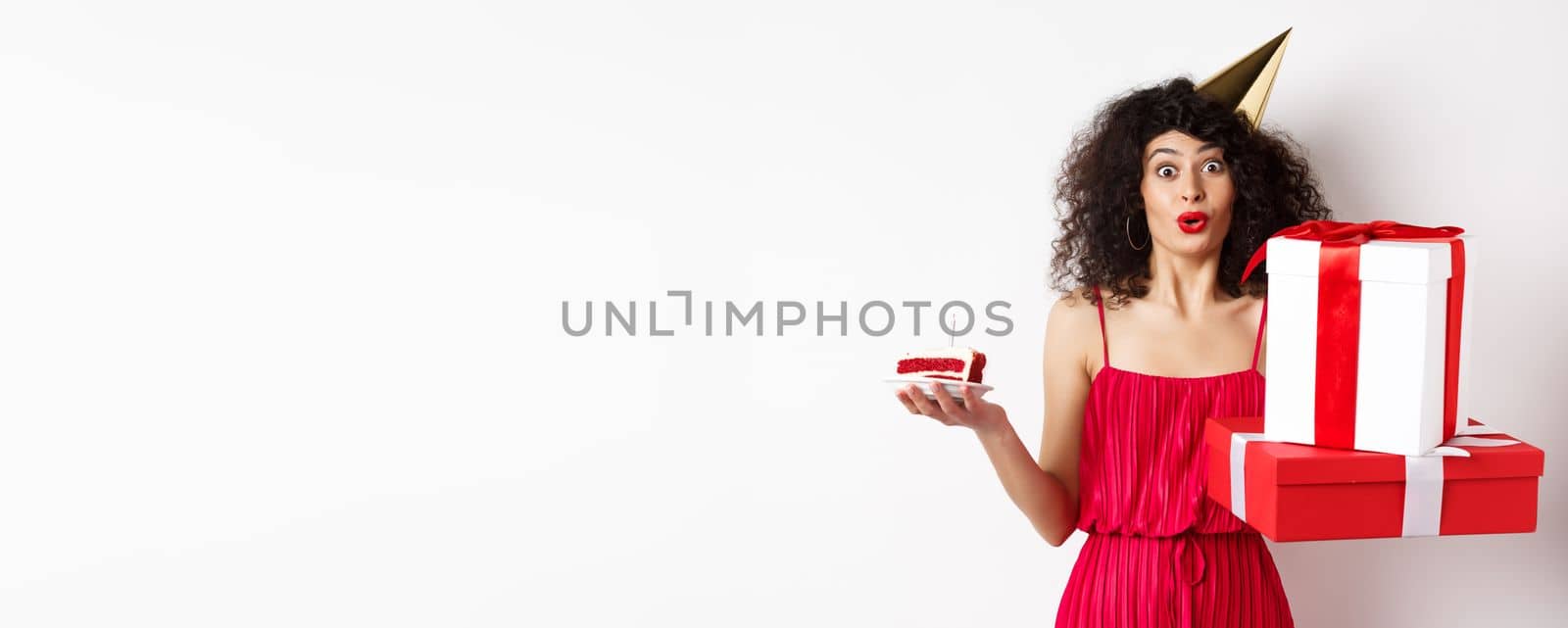 Funny lady in red dress and party hat, celebrating birthday, holding b-day presents and cake with candle, looking amused at camera, standing over white background by Benzoix