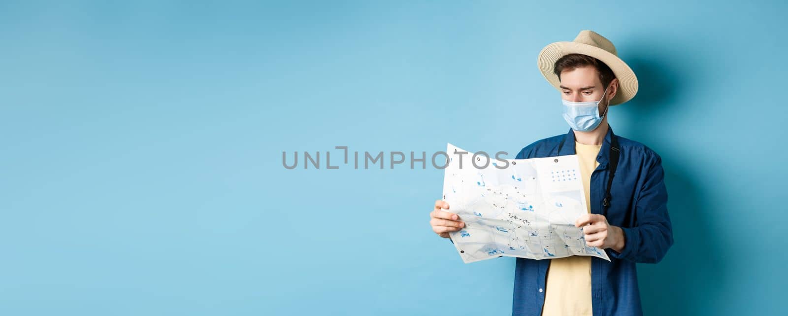 Covid-19, pandemic and travel concept. Tourist looking at map with sightseeing on vacation, wearing summer hat and medical mask from coronavirus, blue background by Benzoix