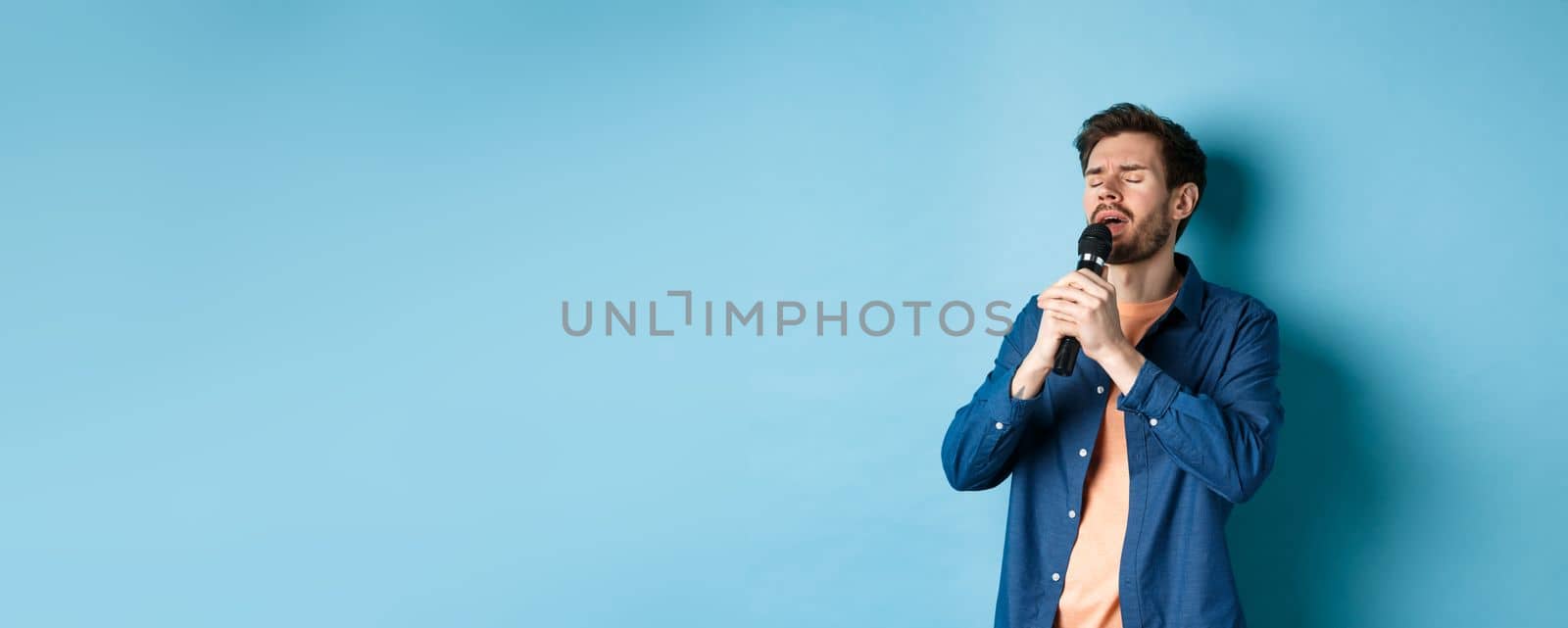 Romantic guy singing in microphone, performing on stage, standing on blue background.