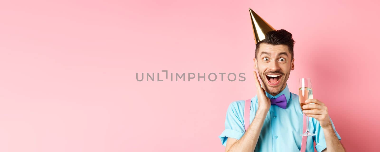 Holidays and celebration concept. Funny young man in birthday hat celebrating, screaming from joy and surprise, raising glass of champagne and smiling, standing on pink background.