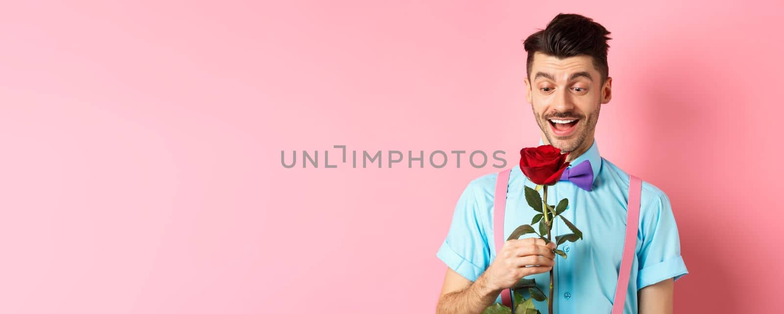 Valentines day and romance concept. Happy boyfriend looking at red rose while waiting for lover in bow-tie, standing on pink romantic background by Benzoix
