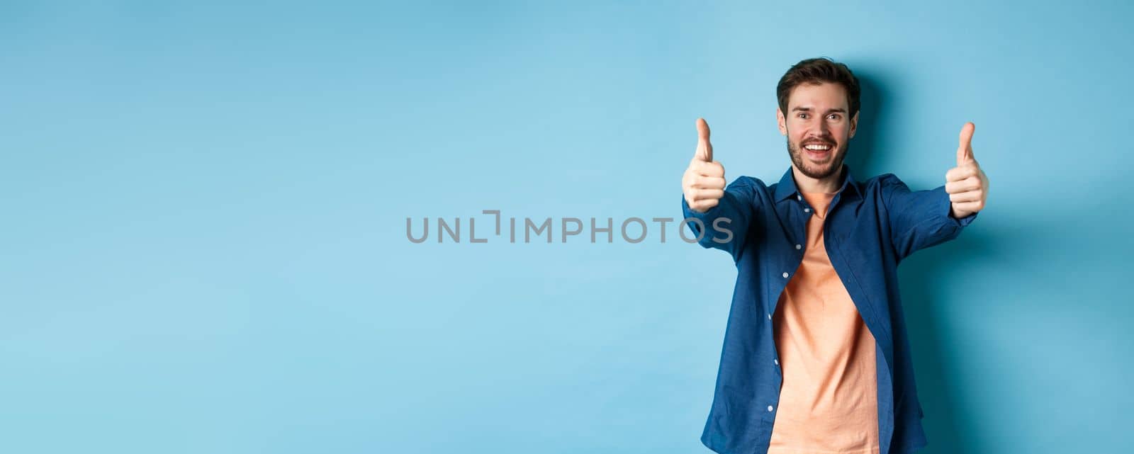 Handsome young man smiling and showing thumbs up in approval, say yes with happy face, standing on blue background.