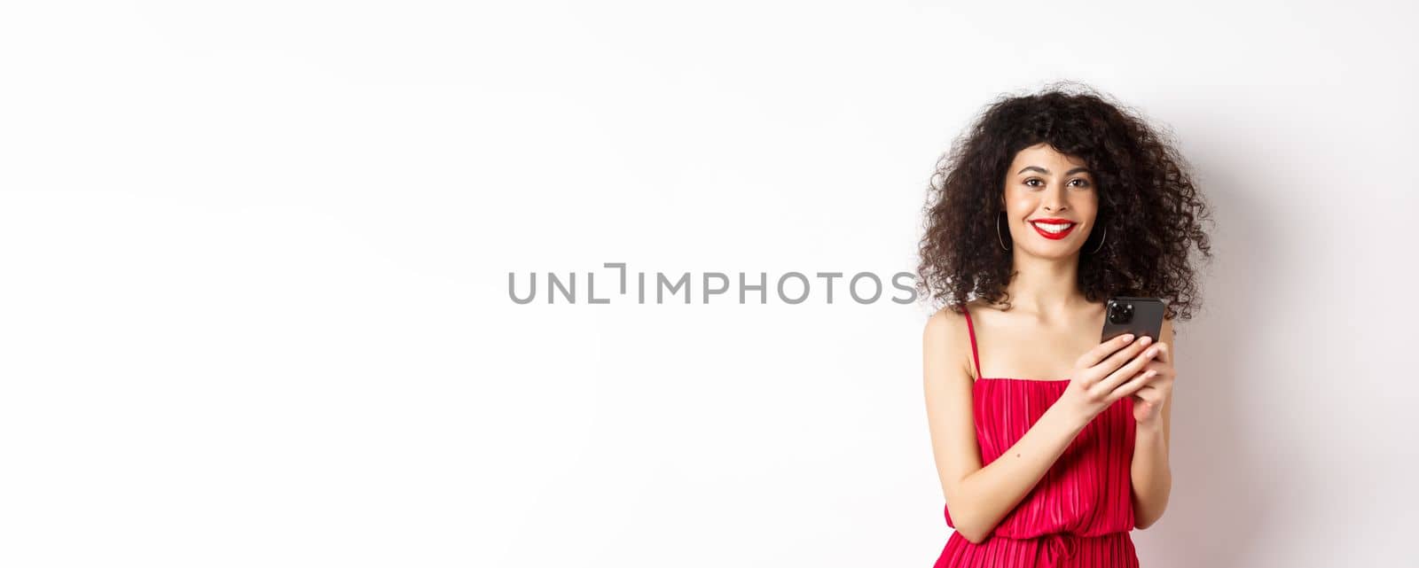 Cheerful woman with curly hair, using smartphone in red dress, smiling at camera, standing over white background. Copy space