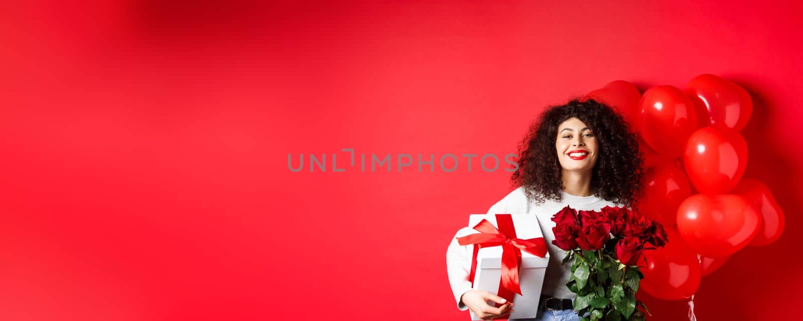 Smiling happy woman holding box with gift and red roses from boyfriend, celebrating Valentines day, standing near romantic hearts balloons, standing over studio background.
