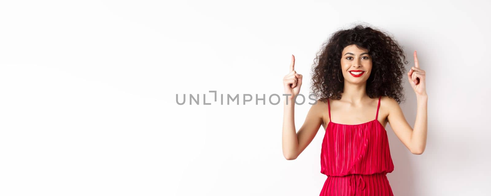 Smiling woman in elegant red dress and makeup, pointing fingers up and showing promo offer on valentines day, standing over white background.