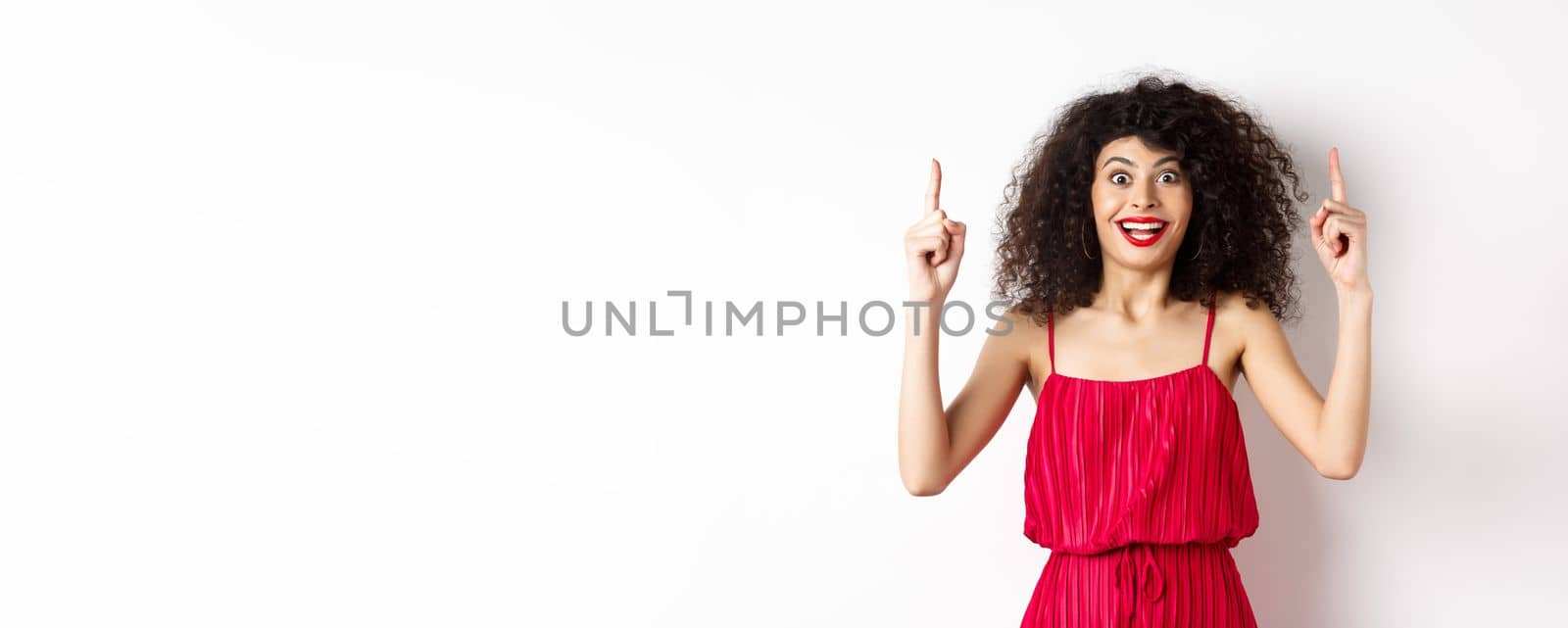 Happy elegant woman in red dress and makeup, smiling amused and pointing fingers up at logo, showing advertisement, standing over white background.