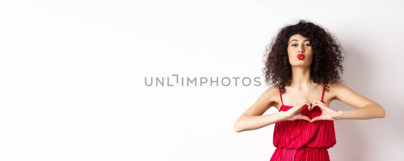 Beautiful lady with curly hair, red dress, showing heart symbol and pucker lips for kiss, love you gesture, standing on white background.