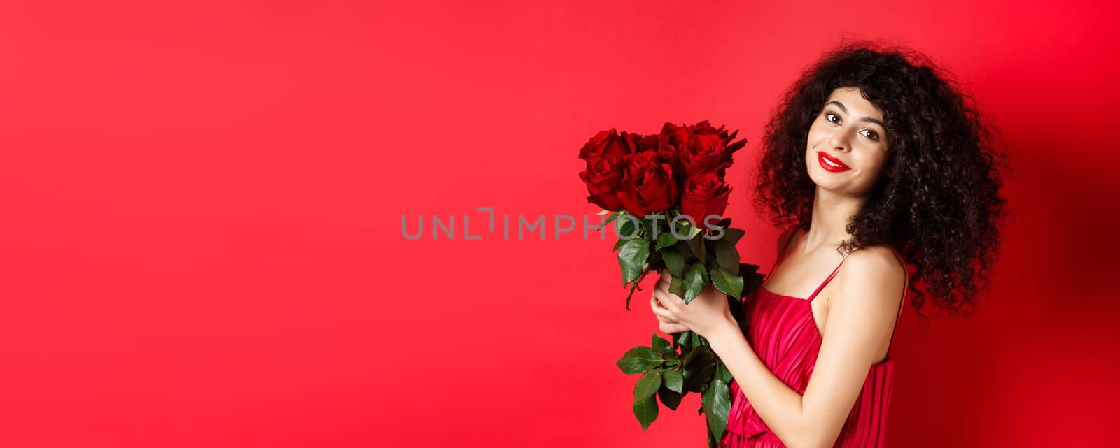 Happy beautiful woman in dress, holding flowers and smiling romantic, standing against red background by Benzoix