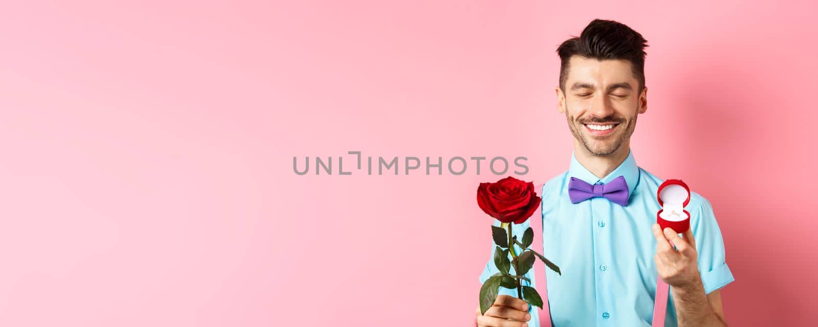 Valentines day. Cute boyfriend making wedding proposal, showing engagement ring in small box and red rose, express love, standing over pink background.