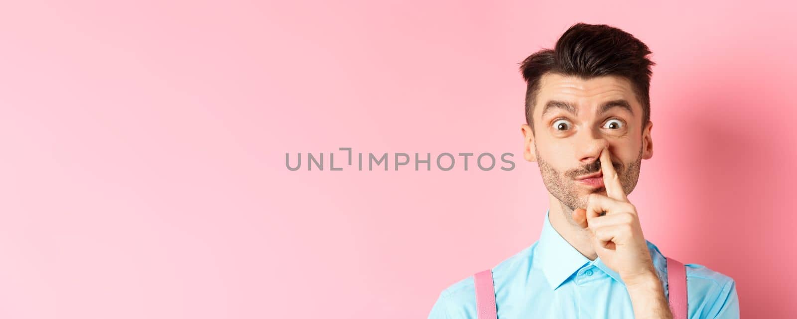 Close-up of funny caucasian man picking nose, staring silly at camera, standing on pink background. Copy space