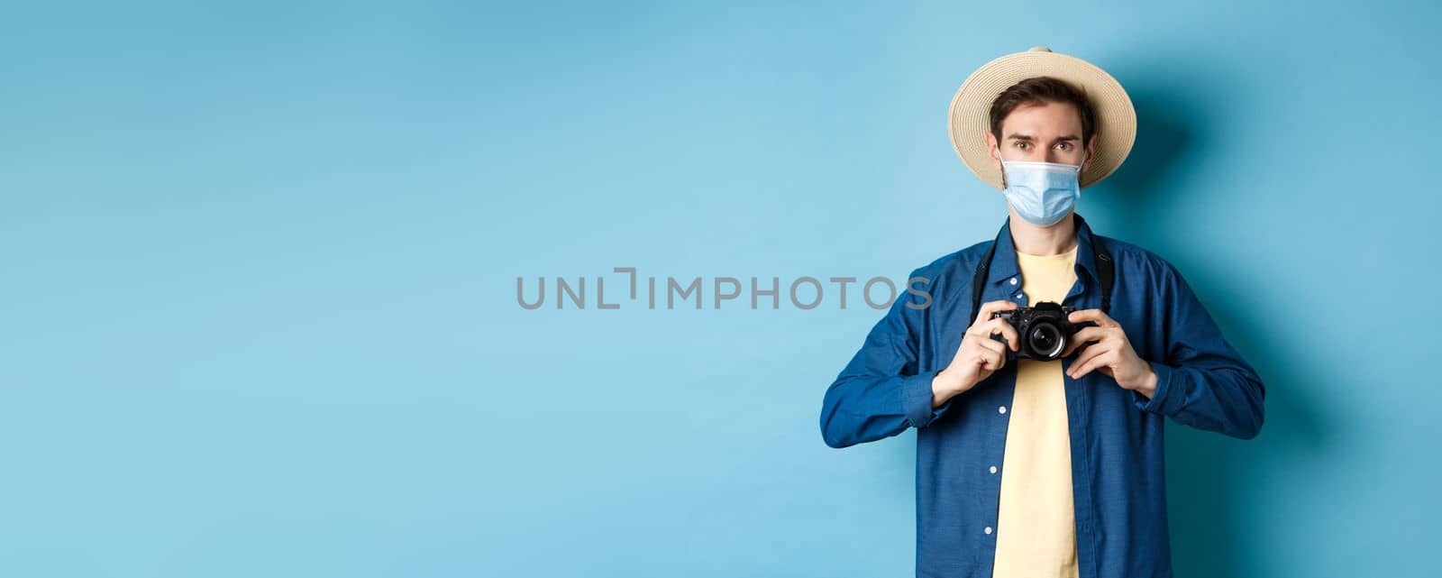 Covid-19, pandemic and travel concept. Cheerful tourist in summer hat and medical mask taking photos, holding camera, standing on blue background.