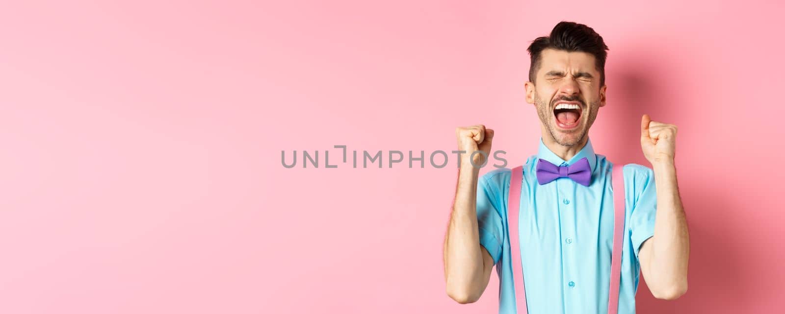 Relieved man shouting from happiness and joy, scream yes with closed eyes and clenched fists, celebrating victory, achieve goal and triumphing, standing over pink background by Benzoix