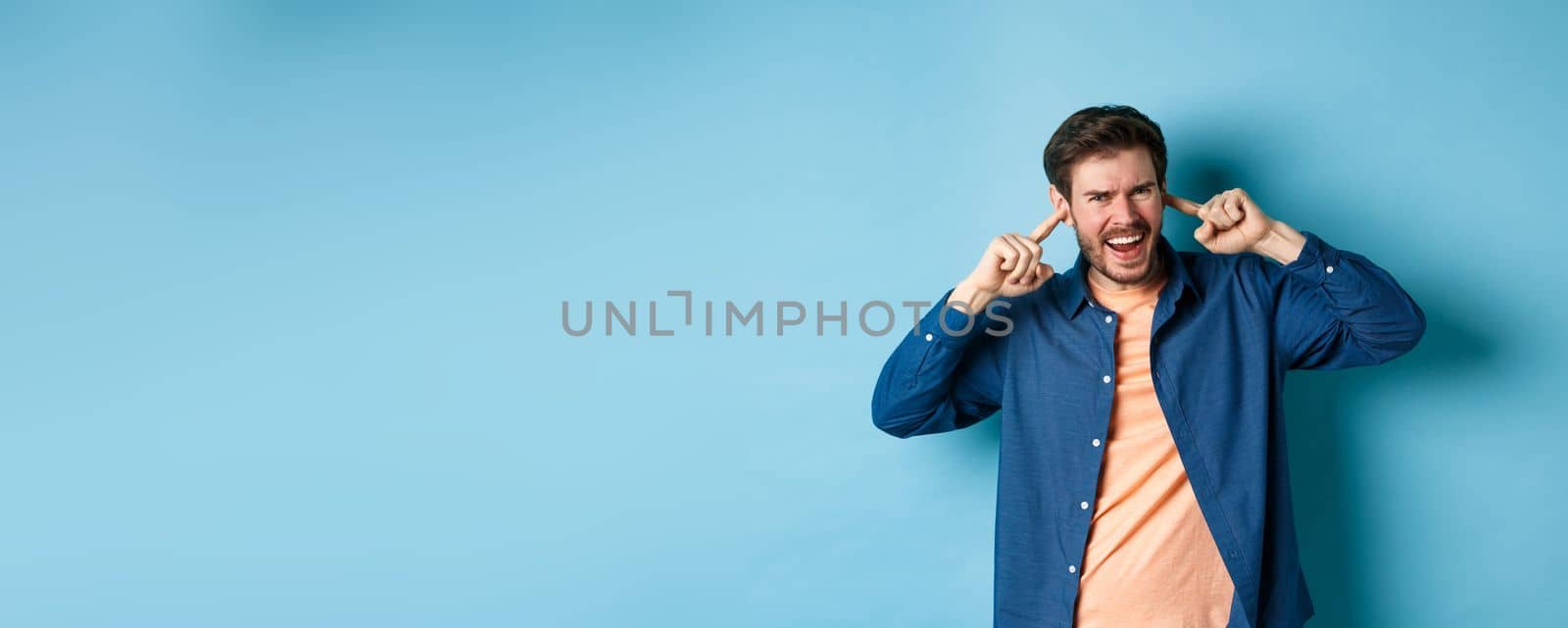 Angry guy shouting with shut ears, block loud music and frowning annoyed, standing on blue background.