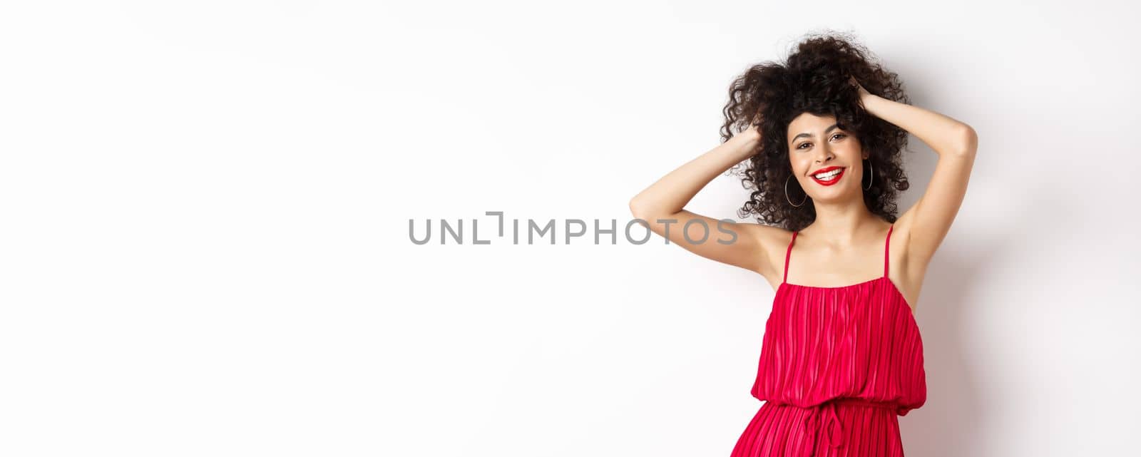 Happy smiling woman touching her curly hair and laughing, standing in red dress on white background by Benzoix