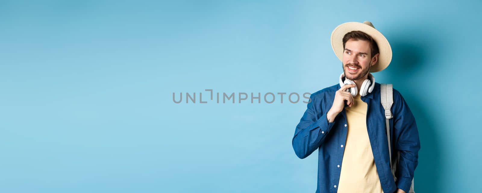 Handsome tourist with backpack and straw hat looking aside, smiling and touching headphones, standing on blue background by Benzoix