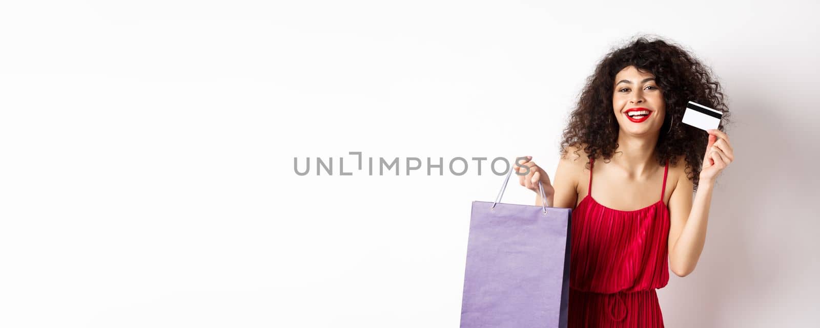 Beautiful woman with curly hair, red dress, showing shopping bag and plastic credit card, white background.