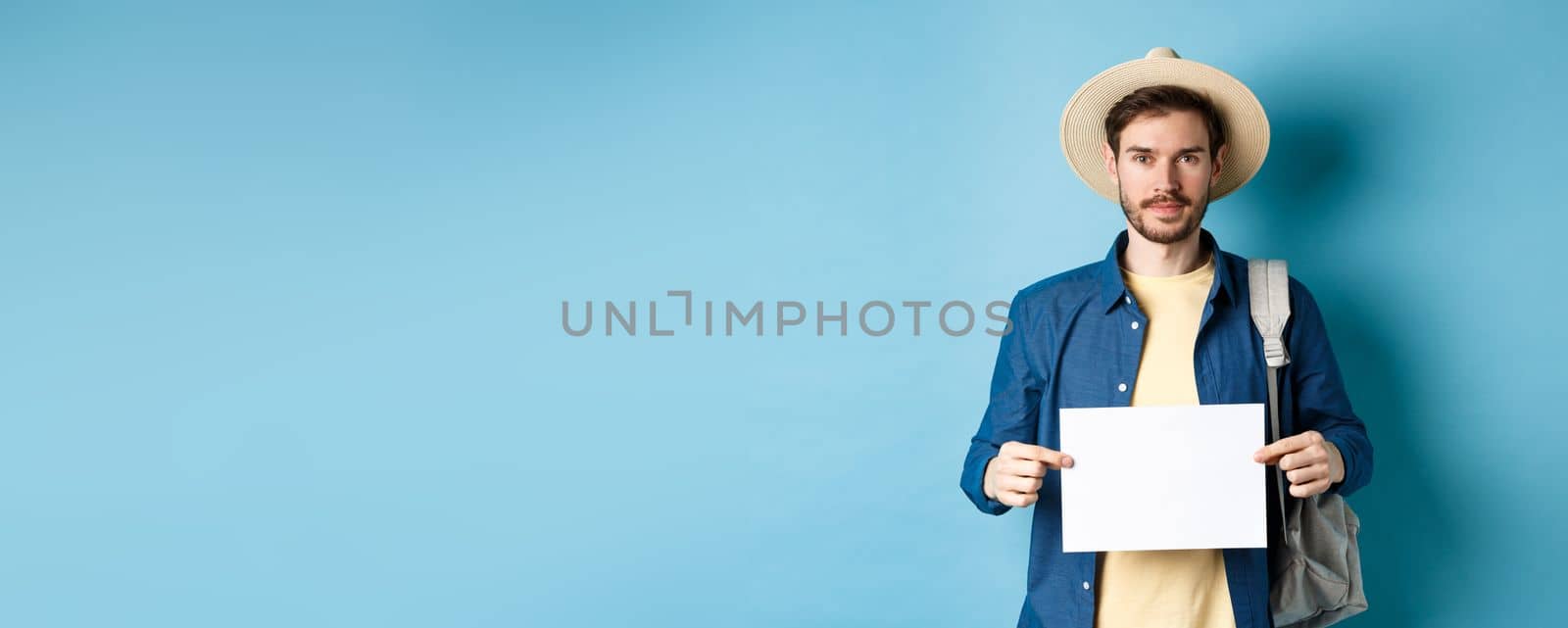 Image of smiling guy in straw hat backpacking, hitchhiking with piece of paper, travelling abroad on summer vacation, blue background by Benzoix