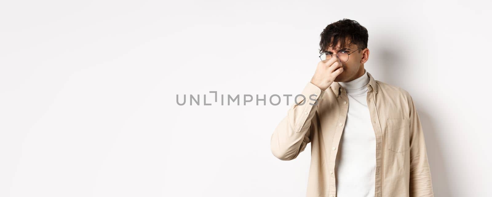 Image of disgusted guy shut his nose from awful smell, looking at something disgusting and stinky, standing on white background.