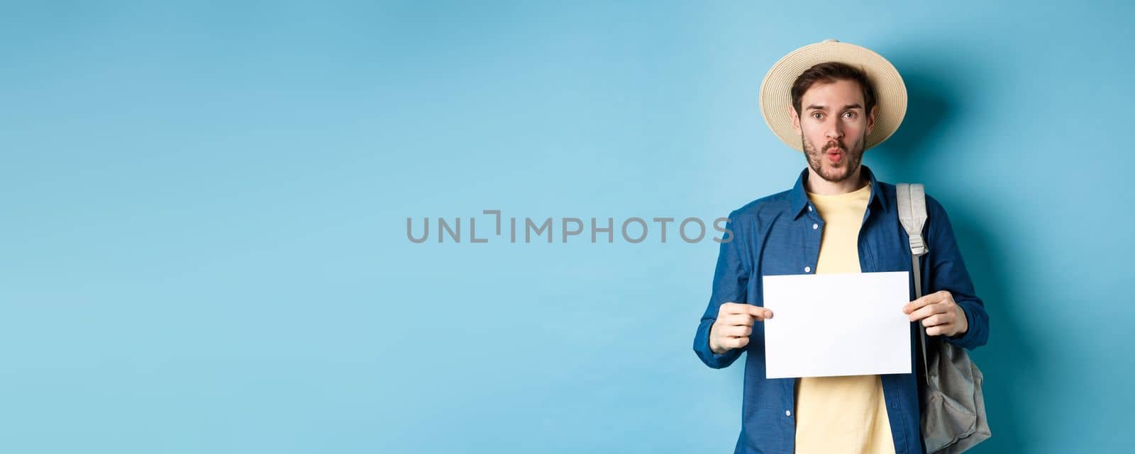 Excited tourist in straw hat, holding empty piece of paper and looking amused, going on summer travel, standing on blue background.