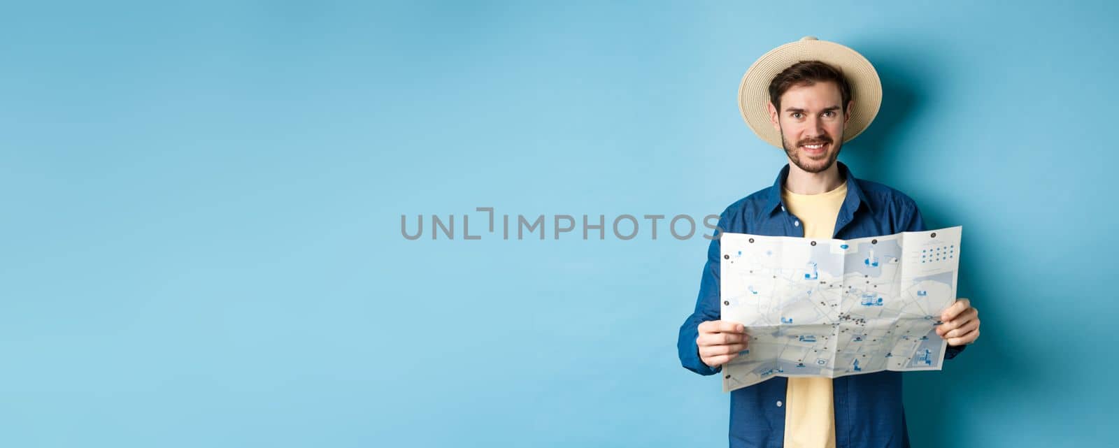 Happy and positive guy on vacation, looking at camera and holding map, smiling excited, going on summer travel, standing on blue background.