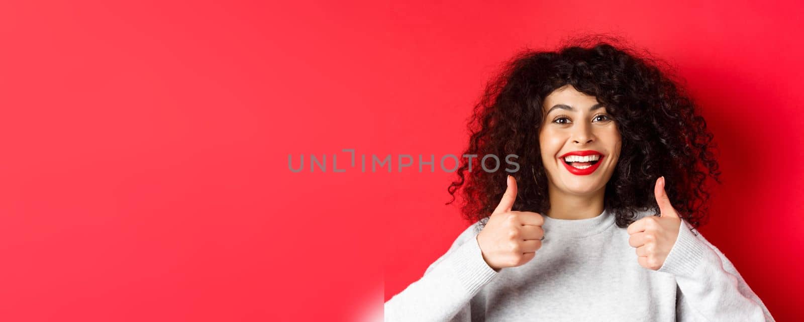 Close up of cheerful caucasian woman showing thumbs up in approval, smiling pleased, like and praise something good, standing on red background.