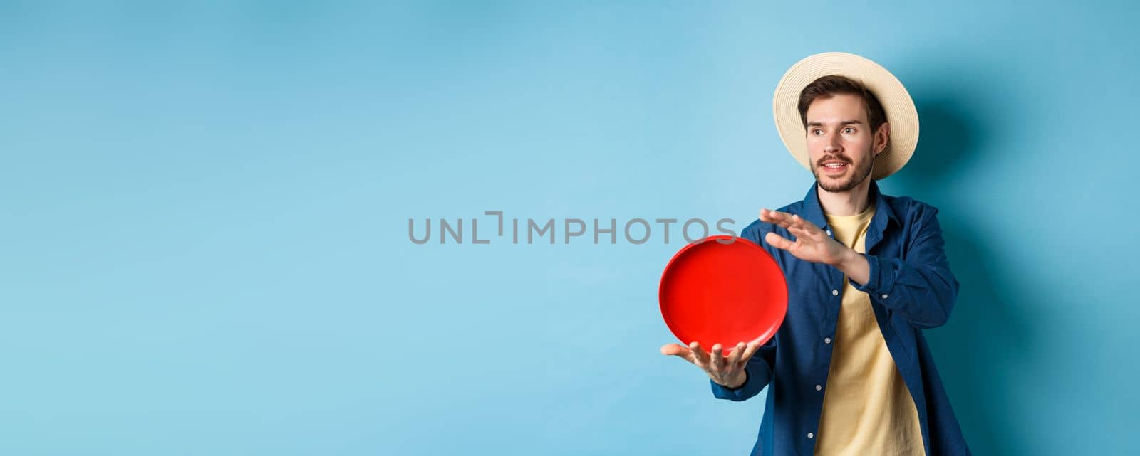 Happy smiling guy throwing frisbee, looking aside at friend, standing on blue background in straw hat by Benzoix