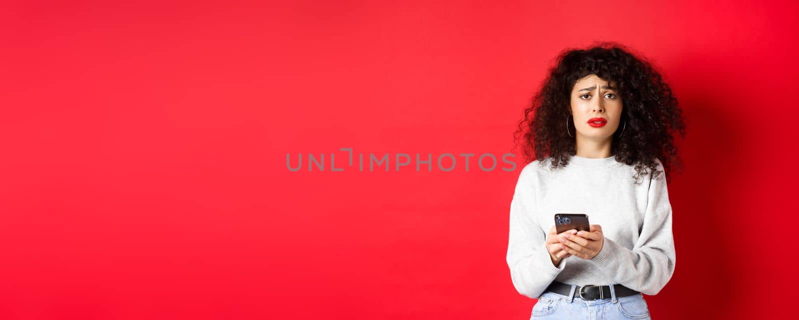 Sad and gloomy woman with curly hair, frowning and feel upset after reading smartphone message, standing disappointed against red background.