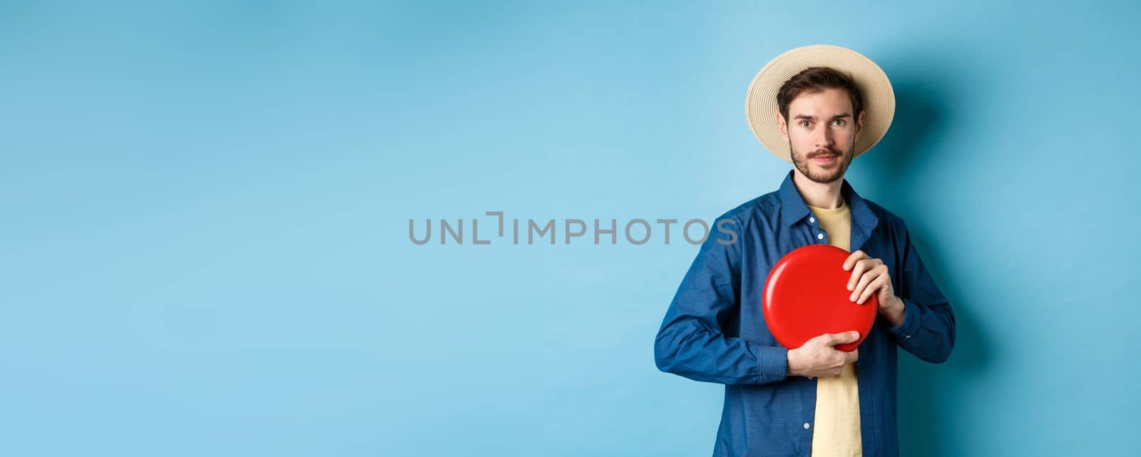 Young man throwing frisbee, playing on summer vacation, standing in straw hat on blue background.