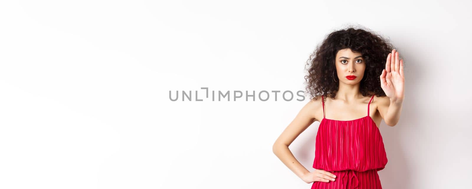 Serious and confident woman in red dress and makeup stretch out hand, tell to stop, prohibit and forbid something, standing over white background by Benzoix