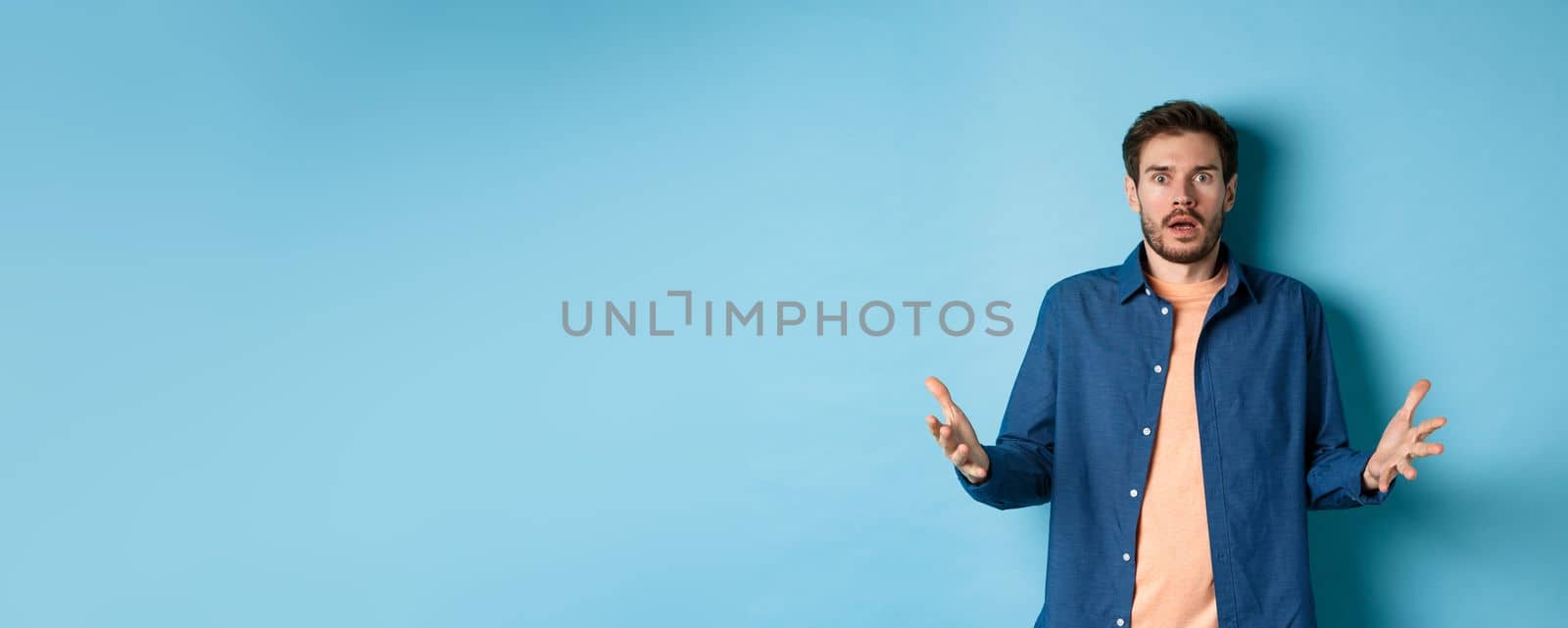 Shocked young man raising hands up and staring at camera startled, look with disbelief, standing on blue background.