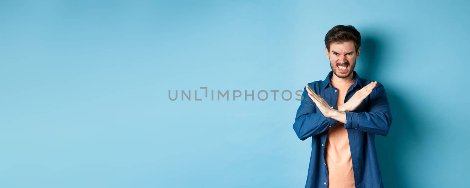 Angry young man frowning and clenching teeth outraged, showing cross gesture to stop or forbid something, standing on blue background.