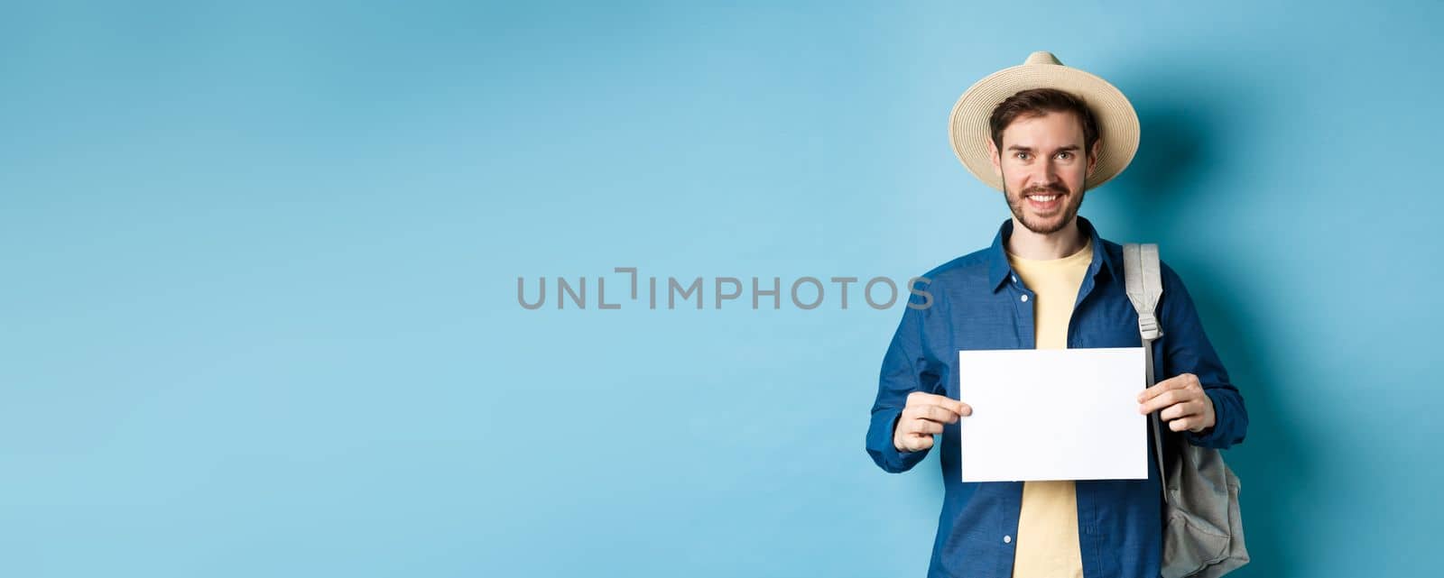 Happy tourist with backpack showing empty piece of paper, smiling at camera, standing on blue background. Concept of summer holiday and vacation.