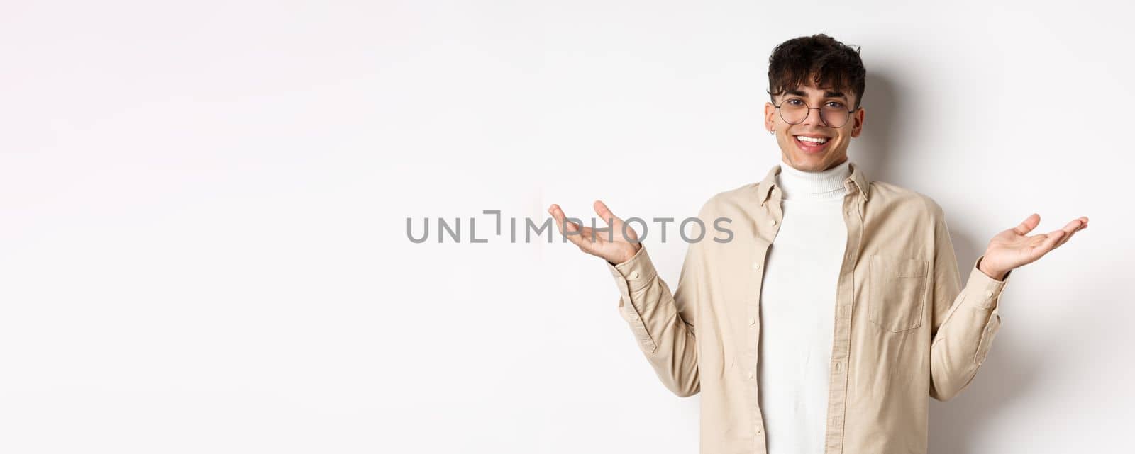 Portrait of happy and pleased young man spread hands sideways and smiling friendly, looking satisfied, standing in glasses on white background.