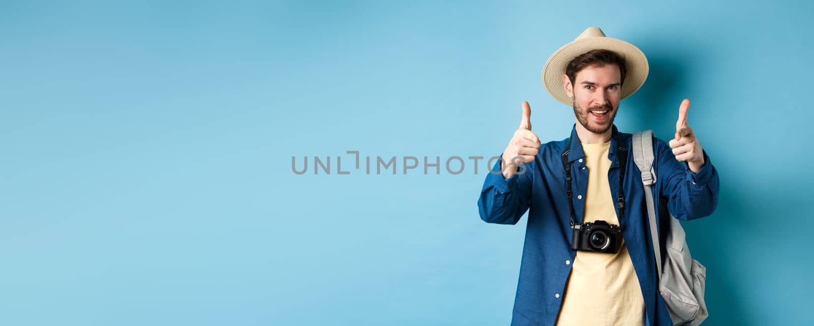 Handsome tourist going on summer vacation with camera and backpack pointing fingers at camera and inviting you, standing on blue background.