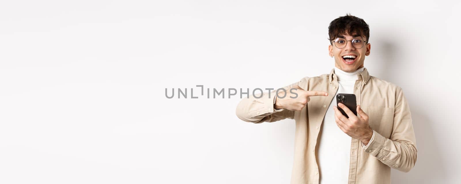 Technology and online shopping concept. Happy hipster guy pointing finger at smartphone screen and smiling, checking out app promo, standing on white background.