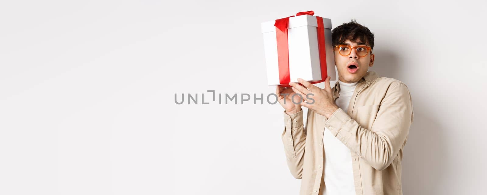 Holidays concept. Funny young man in glasses shaking gift box, listening what inside present and gasping amazed, standing on white background.