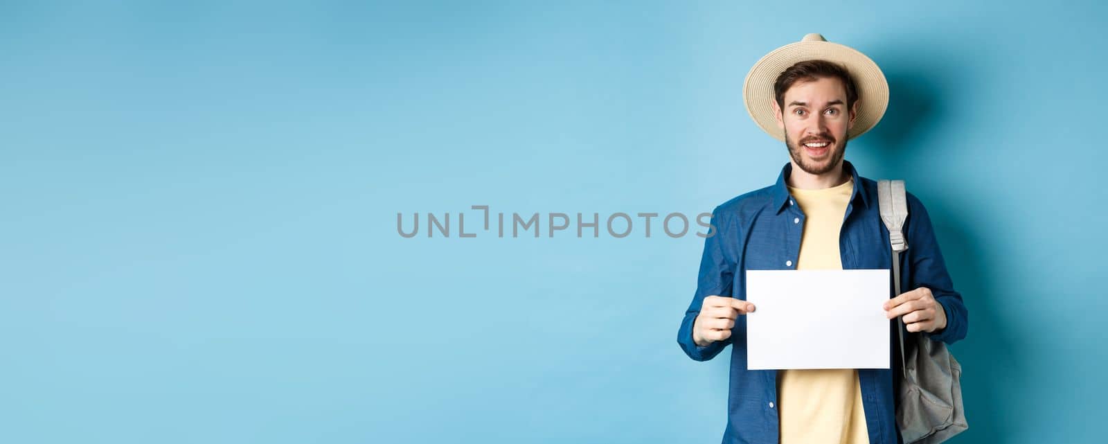 Cheerful tourist in summer hat, showing empty piece of paper and smiling, hitchhiking with backpack, standing on blue background by Benzoix