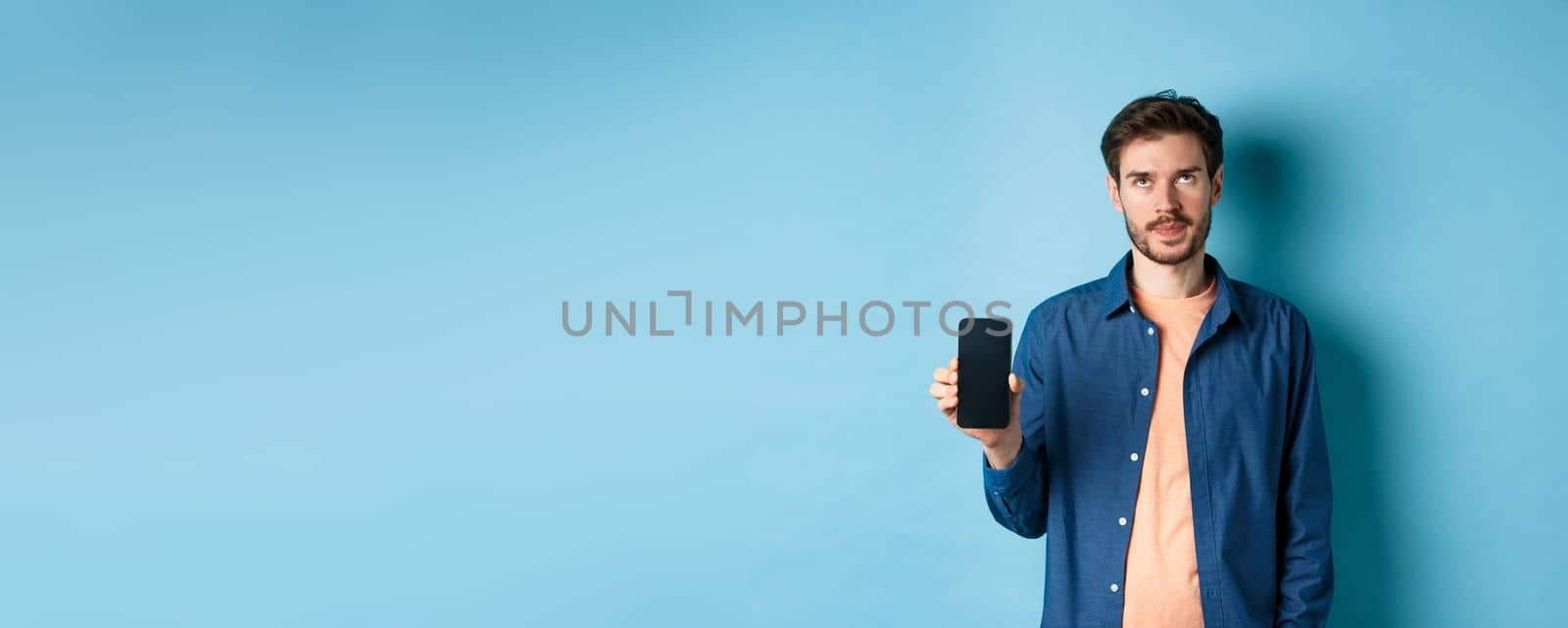 Annoyed young guy showing empty smartphone screen and roll eyes up bothered, standing on blue background.