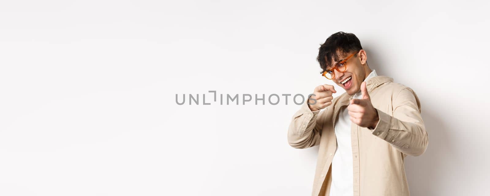 Handsome funny man in glasses smiling, pointing fingers at camera, choosing or inviting you, standing on white background.