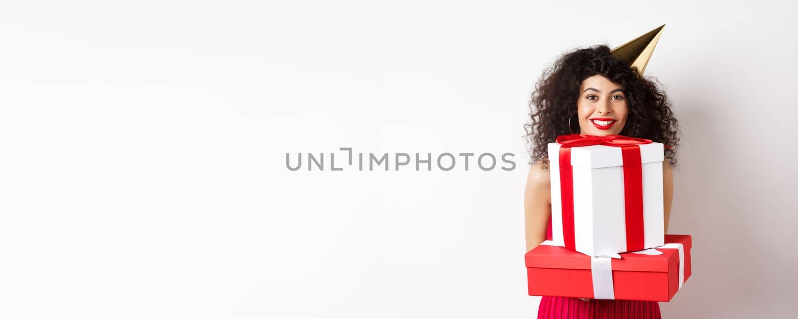 Holidays and celebration. Beautiful lady with curly hair, wearing party hat and holding big presents, smiling happy, receiving birthday gifts, standing on white background by Benzoix