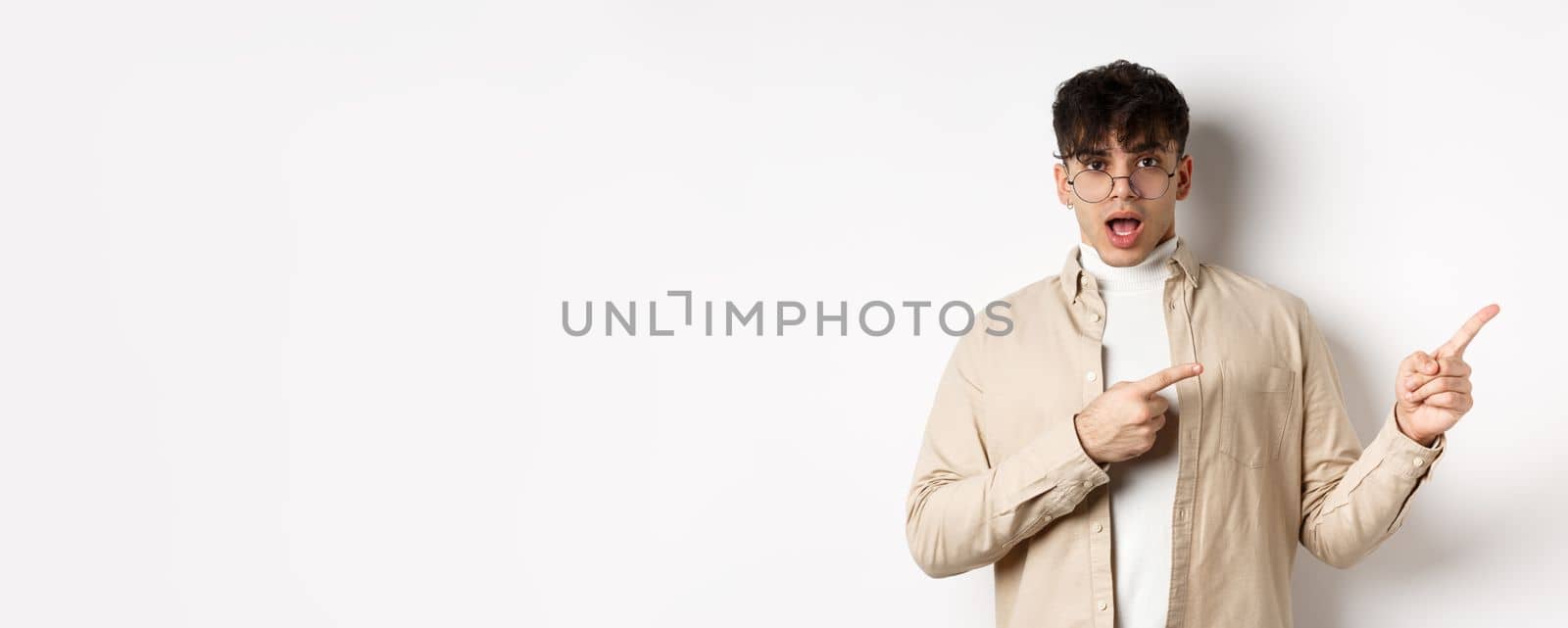 Shocked young man in glasses pointing fingers right at empty space, asking about advertisement, standing on white background.