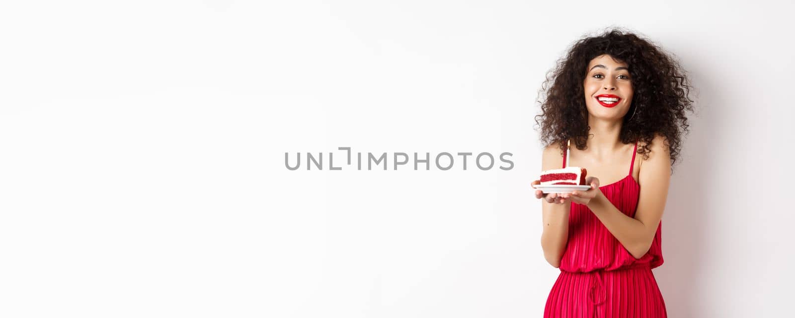 Beautiful lady in red dress celebrating birthday, holding piece of cake with candle and smiling, standing happy on white background.